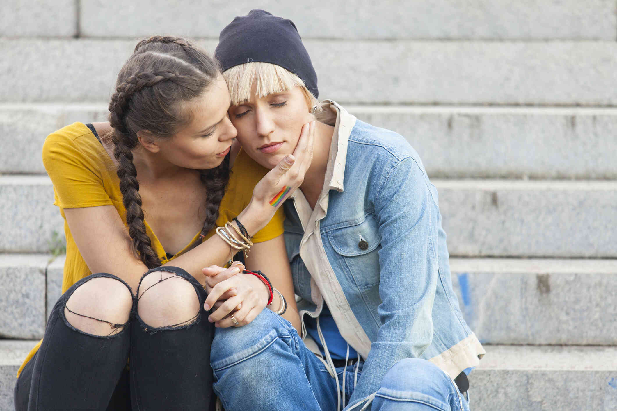 A female couple sit on cement steps outside and lean into each other comfortingly with serious expressions while holding hands.